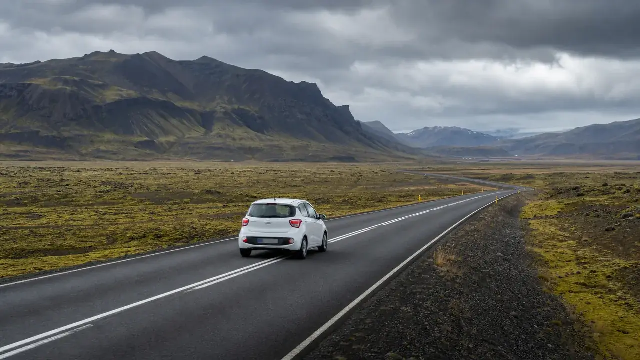 A small car traveling on a paved road through a remote Icelandic landscape beneath an overcast sky.